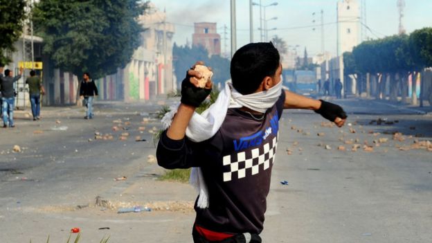 Um jovem atira uma pedra em uma rua cheia de fumaça