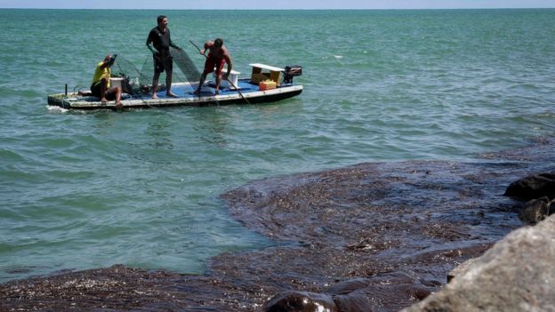 Jangada com três homens segurando rede de pescar e mar com manchas de óleo, no litoral do Pernambuco