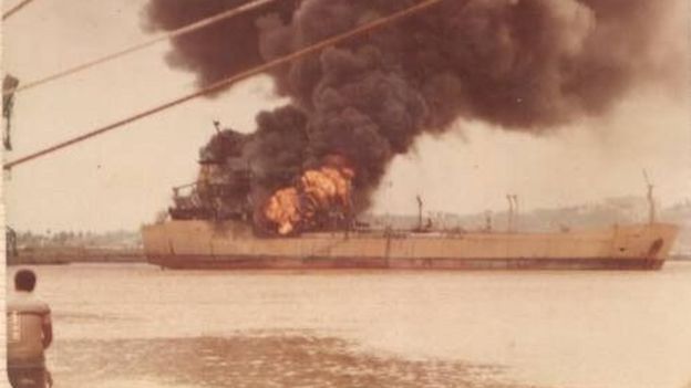 Foto antiga, mas colorida, mostra homem na beira do mar, com navio pegando fogo ao fundo