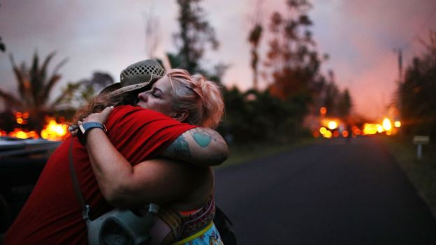 Duas pessoas se consolam em um abraço, com a erupção do vulcão visível ao fundo