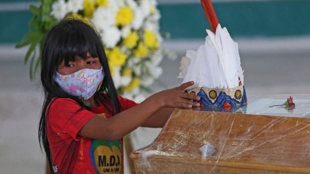 Indigenous child wearing a mask puts headdress on coffin in Brazil