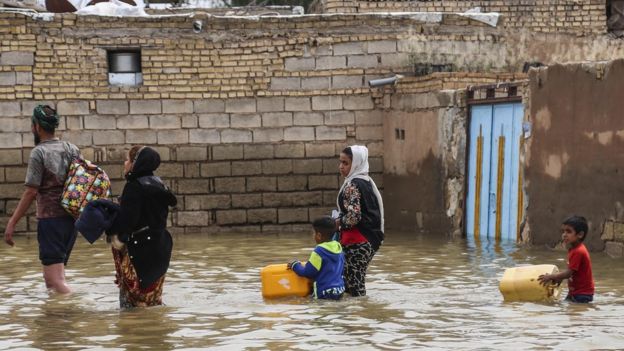 A family wade through the water outside destroyed buildings carrying blankets and bottles
