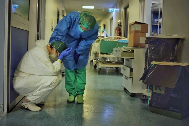 Em corredor de hospital, um membro da equipe consola outro, ajoelhado com as mãos na cabeça