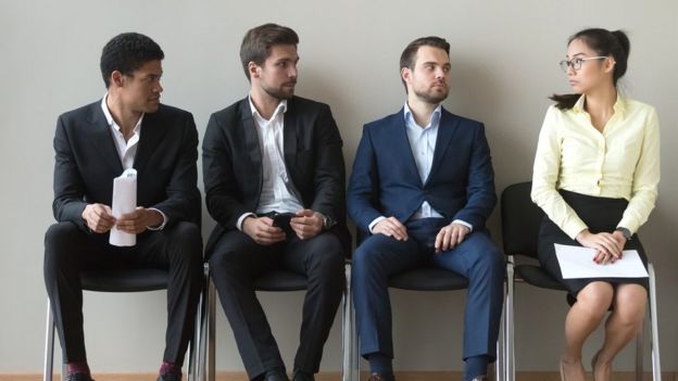 Diverse male applicants looking at female rival among men waiting for at job interview,