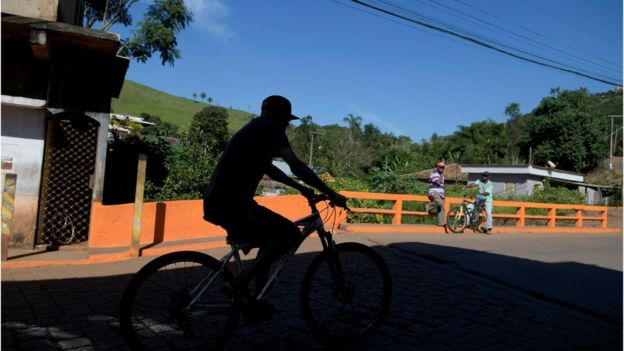 Sombra de homem passando de bicicleta em ponte de Barão de Cocais, durante dia ensolarado e observado por outros dois homens