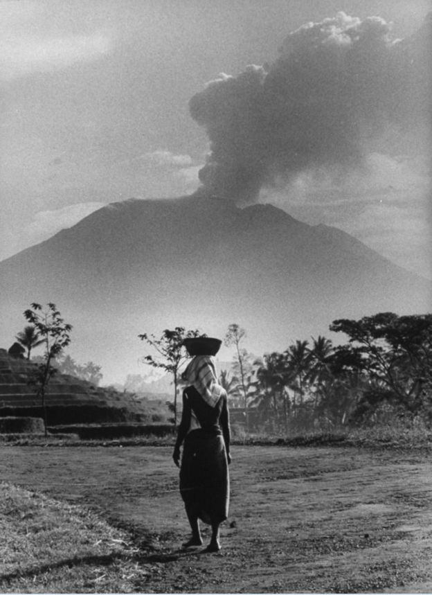 Seorang perempuan Bali mengangkut kebutuhan dasar di kepalanya, dengan Gunung Agung yang masih mengepulkan asap di kejauhan pada tahun 1963.