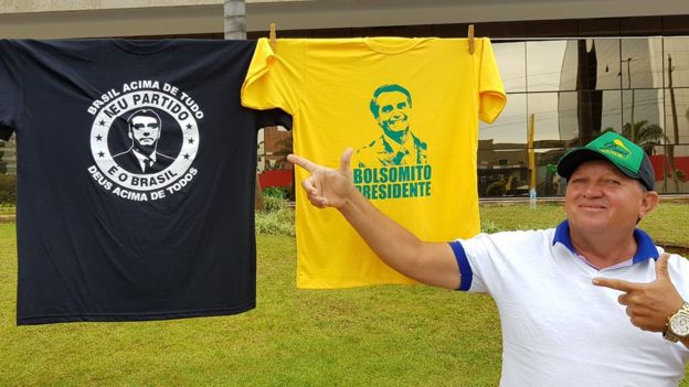 Ambulante mostra camisetas em homenagem a Bolsonaro à venda, fazendo sinal de arma