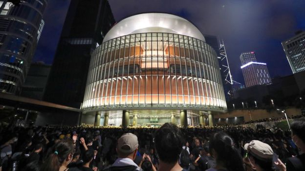 An illuminated modern building of steel and glass, circular at the front, is seen against a dark sky, with innumerable people watching it passively from a distance