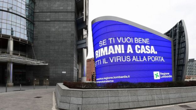A display reads "Stay home, leave the virus at the door" on Piazza Gae Aulenti in Milan on March 12, 2020, as Italy shut all stores except for pharmacies and food shops in a desperate bid to halt the spread of a coronavirus that has killed 827 in the the country in just over two weeks. (Photo by Miguel MEDINA / AFP) (Photo by MIGUEL MEDINA/AFP via Getty Images)