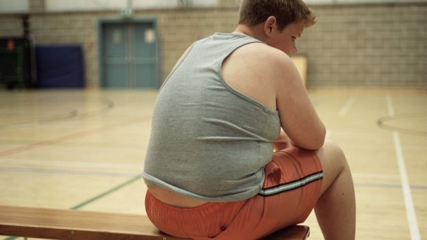 Garoto obeso sentado em banco em ginásio de colégio