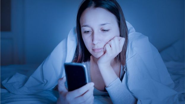 Chica mirando un celular en su cama, de noche.