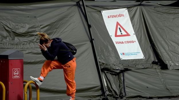 Mulher com a máscara abaixada apoia as mãos na cabeça do lado de fora de uma tenda onde está escrito "atenção, zona covid+"