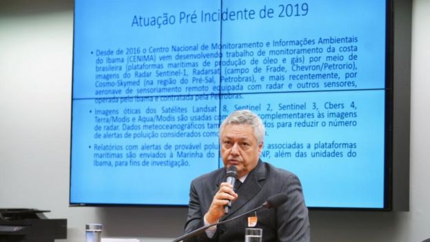 Pedro Alberto Bignelli aparece sentado, falando em microfone e com tela com apresentação projetada atrás