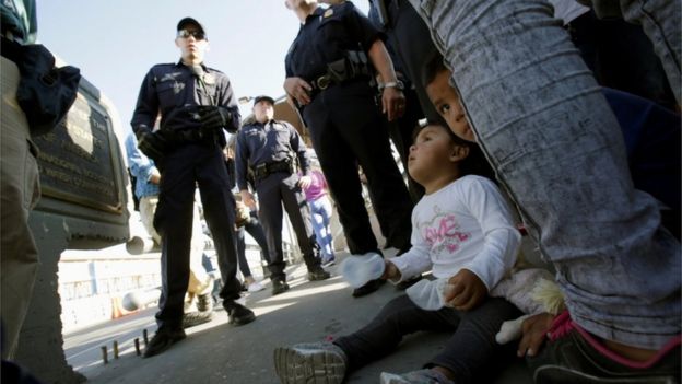 Photo showing children sitting at parents feet in front of border agent officials