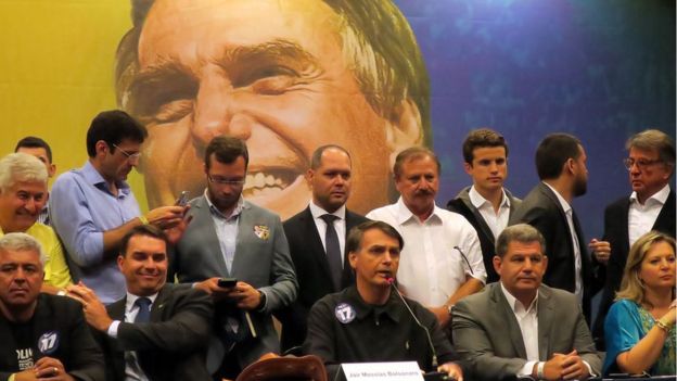 Bolsonaro em coletiva de imprensa quando era candidato, em 2018, cercado por correligionários e em frente a um grande painel com sua foto exposta