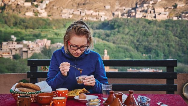 Mulher come à mesa de café da manhã; no plano de fundo, montanha com casas em paisagem marroquina