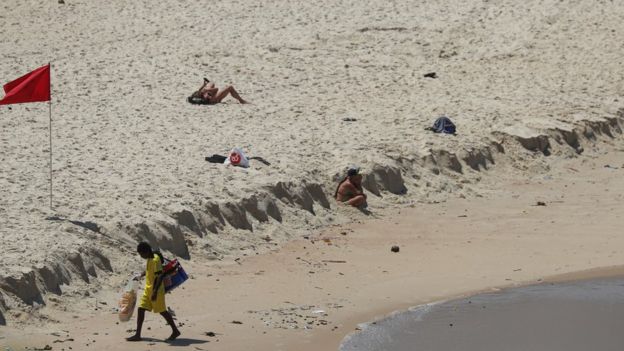 Ambulante caminha na area de praia no Rio de Janeiro, com duas mulheres pegando sol ao fundo