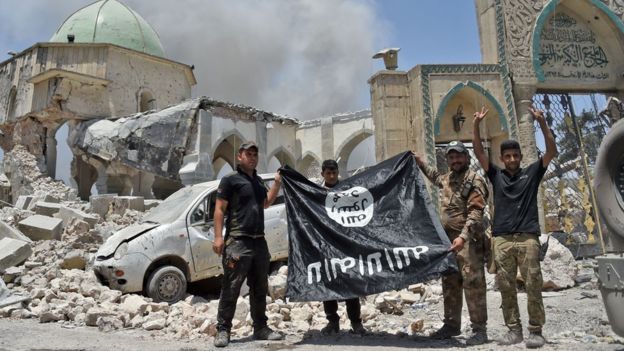 Soldados iraquíes sostienen una bandera de EI al revés, frente a las ruinas de una mezquita, junio de 2017