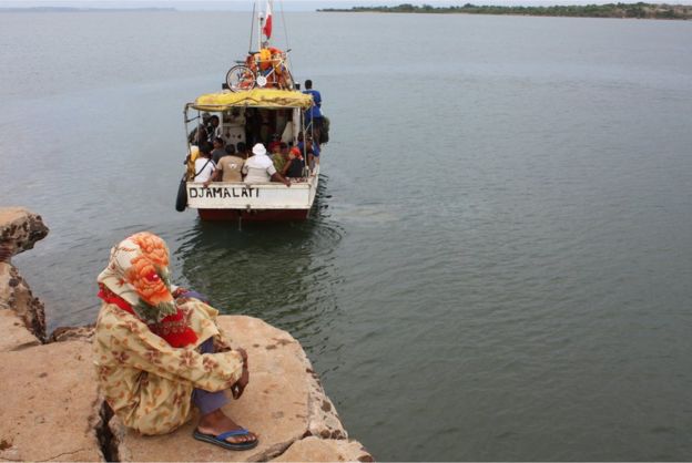 A woman sits on the shore close to a small motorboat - known as a kwassa-kwassa - which is loaded with passengers