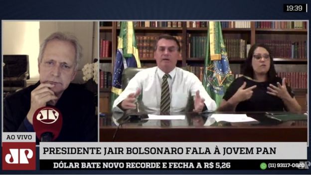 Tela de TV mostra, em um quadro, entrevistador; no outro, Bolsonaro falando ao lado de intérprete de Libras