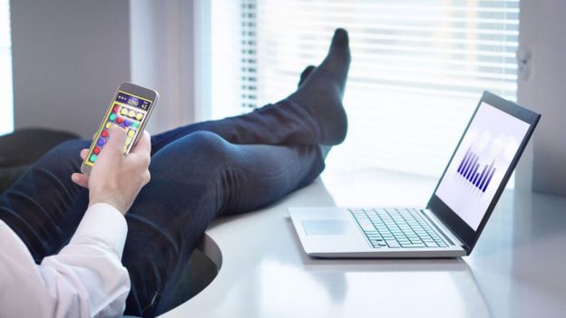 Un hombre juega un videojuego en su celular con los pies sobre el escritorio.