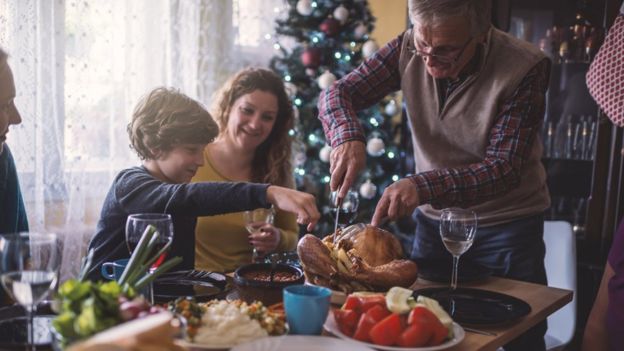 Família come ceia de Natal ao redor de uma mesa, com árvore decorada ao fundo