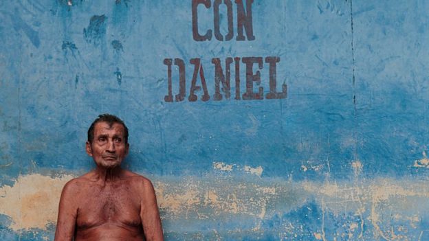 Un hombre sin camisa frente a una pared donde se lee 