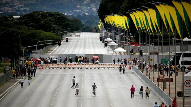 Vista ampla de pista em Brasília com pontos de bloqueio, alguns pedestres e bandeiras em verde e amarelo