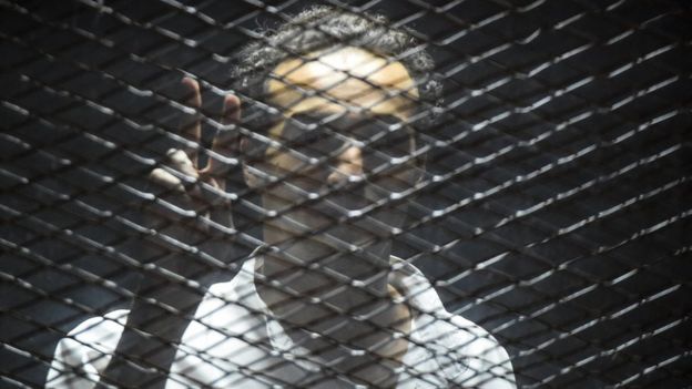 Egyptian photographer Mahmoud Abdel Shakour Abou Zeid, also known as Shawkan, flashes the victory gesture from inside a soundproof glass dock, during his trial in the capital Cairo on July 28, 2018