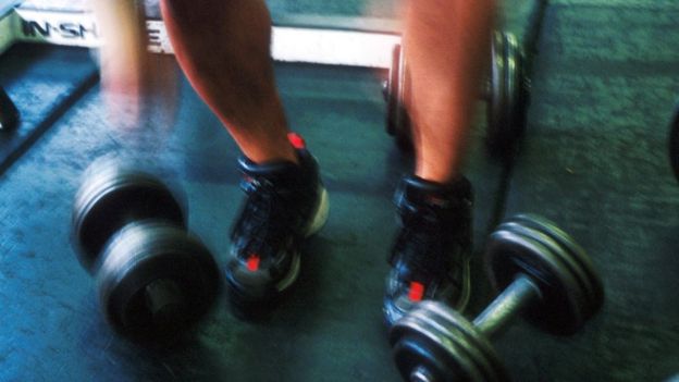 Imagem mostra halteres de musculação e pernas de uma pessoa com tênis esportivo