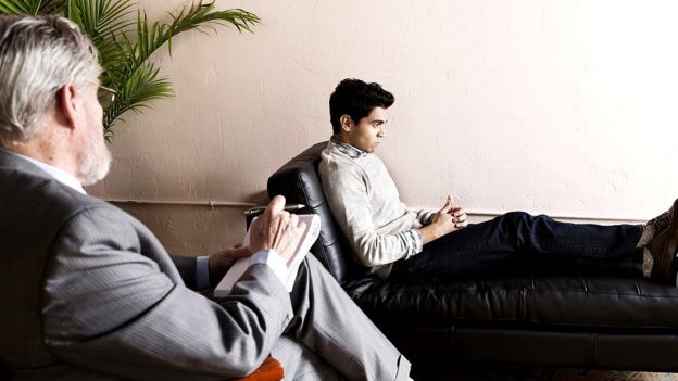 Un hombre joven en un diván con un psicólogo de espaldas.