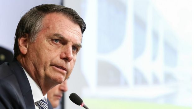 Bolsonaro fala no microfone em posição de perfil, com imagem da fachada do Palácio do Planalto ao fundo