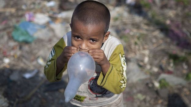 Criança usando camisinha como balão de ar, no meio do lixo