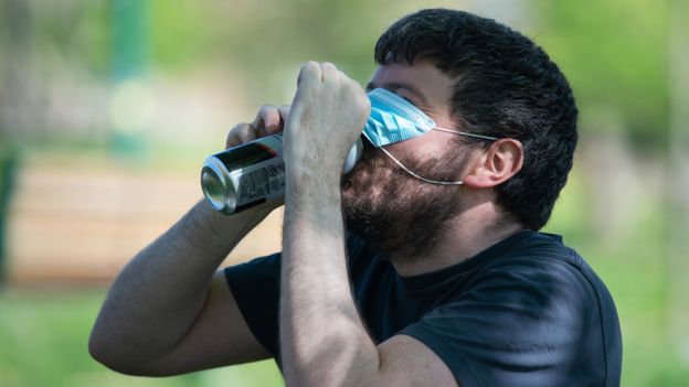 Man lifts a face mask to swig lager from a can