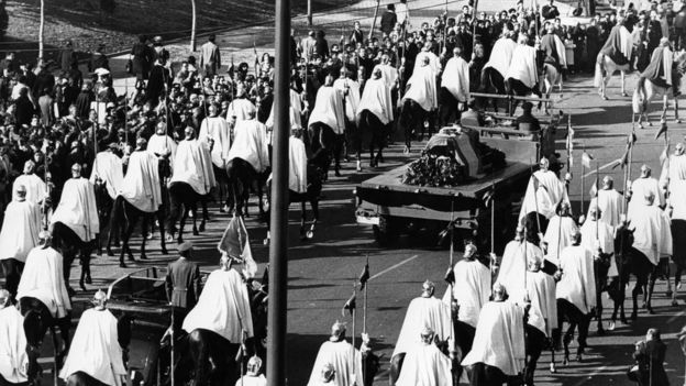 The funeral procession for Franco in 1975 towards the Valley of the Fallen. His flag-draped coffin is flanked by men wearing white cloaks on horseback