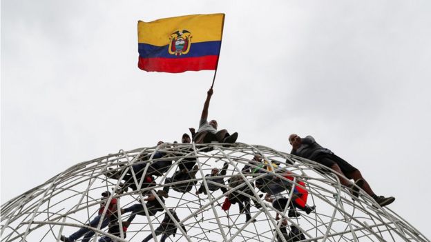 Manifestantes trepados en una escultura, con la bandera de Ecuador.