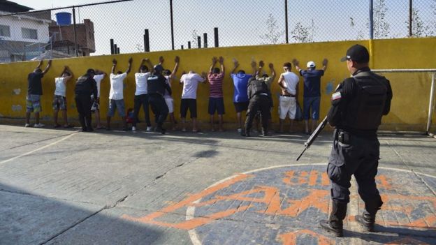 Policial da Faes diante de homens parados contra um muro