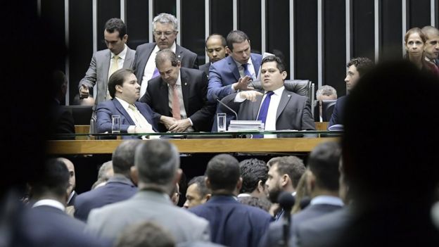 Ao centro, Rodrigo Maia e Davi Alcolumbre aparecem sentados em mesa no Plenário da Câmara, rodeados por parlamentares e assessores que falam com eles