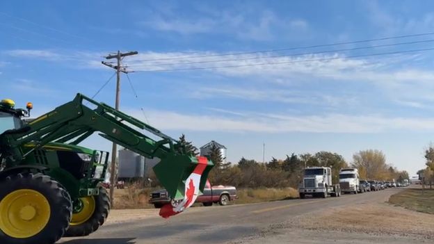 Green tractor with Canadian flag on road in front of line of trucks