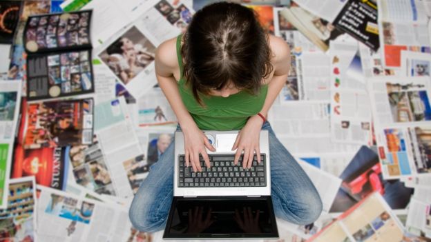 Mulher sentada no chão usando laptop rodeada por revistas e jornais