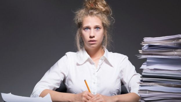 Una mujer con rostro de estrés y una pila de papeles a su izquierda