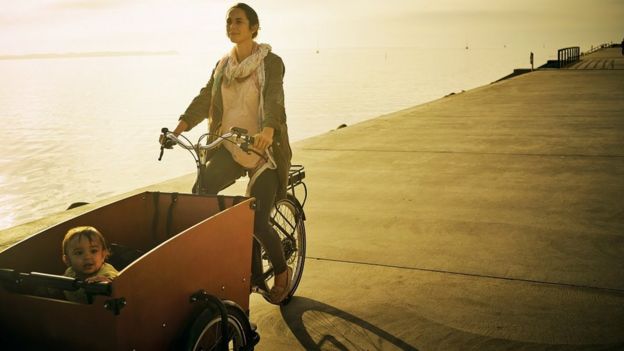 Una joven madre pedalea una bicicleta con su hijo en un carrito en frente