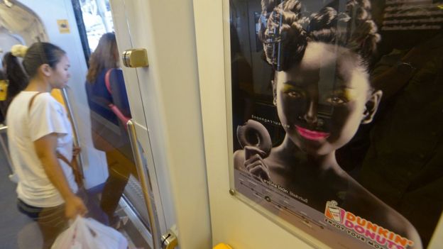 Thai passengers walk past a Dunkin' Donuts advertising campaign featuring a woman with black face make-up displayed at a skytrain station in Bangkok on September 3, 2013