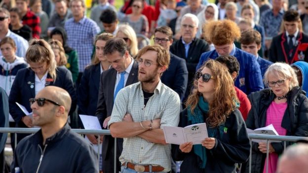 A crowd of people watch the ceremony, some with heads bowed, some reading from service sheets, in Hagley Park on March 29