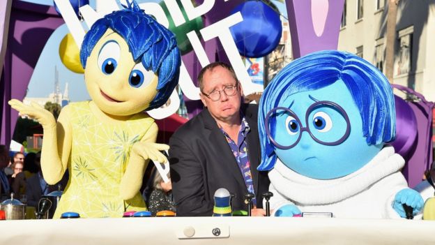 John Lasseter is pictured comically frowning in exaggerated sadness, seated between the characters of Joy and Sadness from Pixar's 