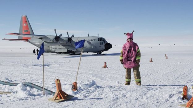 A woman in a whoopee cushion costume stands on a frozen airfield in front of a plane