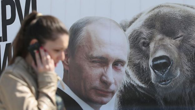 Una mujer con un celular frente a un retrato de Vladimir Putin