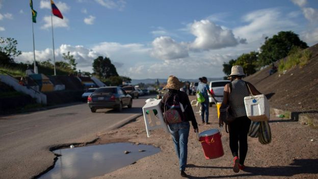 Imigrantes caminham carregando malas e bancos no acostamento de uma estrada na fronteira entre Brasil e Venezuela