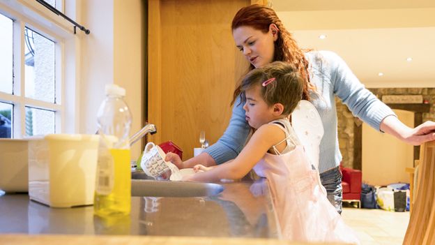 Imagen genérica de una niña trans, vestida de hada, lavando los platos con su madre.