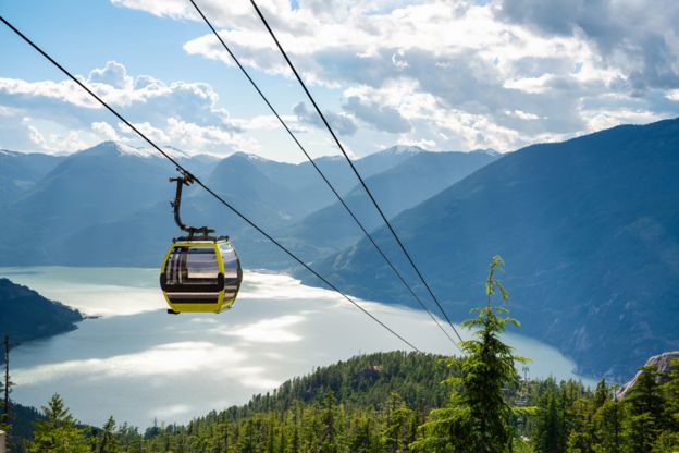A shot shows a cable car move down towards a stunning lake at the base of rolling green mountains carpeted thickly with pine trees